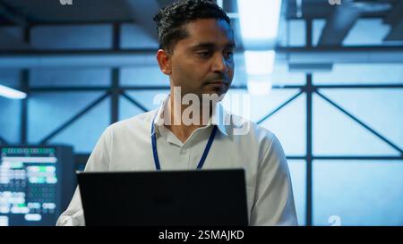 System administrator in data center using notebook to design, implement and manage gear, ensuring optimal performance and reliability. Indian man in server room coding on laptop, camera A Stock Photo