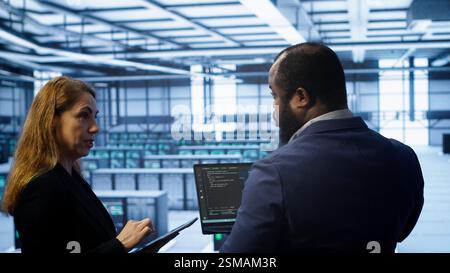 Teamworking coworkers doing maintenance using devices, prolonging data center hardware life span. Colleagues doing brainstorming in server hub, monitoring server infrastructure using devices, camera A Stock Photo