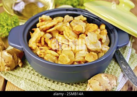 Jerusalem artichokes roasted in a roasting pan with a lid, fresh tubers of on a napkin, parsley, vegetable oil on a wooden boards background Stock Photo