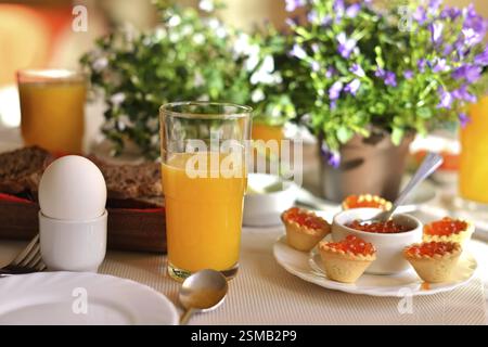 Festive continental breakfast with red caviar, soft-boiled egg and orange juice. Table decorated with a bouquet of bells flowers. Sunny light Stock Photo