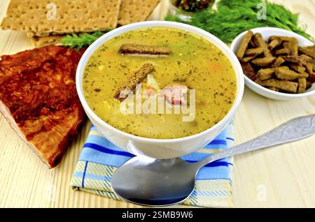 Pea soup in a bowl, rye crackers on a plate, spoon, pepper, crispbread, cloth, bacon, dill against a wooden board Stock Photo