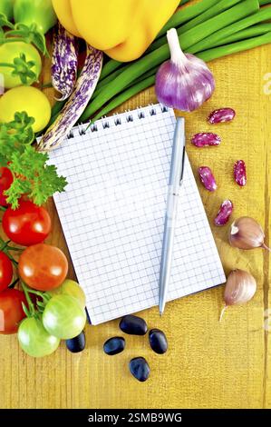 Notebook, silver metallic pen, small twigs of tomatoes, yellow bell pepper, green onions, parsley, whole and two cloves garlic, black beans and kidney Stock Photo