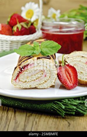 Biscuit roulade with cream and jam, a jar of jam, napkin, chamomile flowers, strawberries, mint on the background of wooden boards Stock Photo