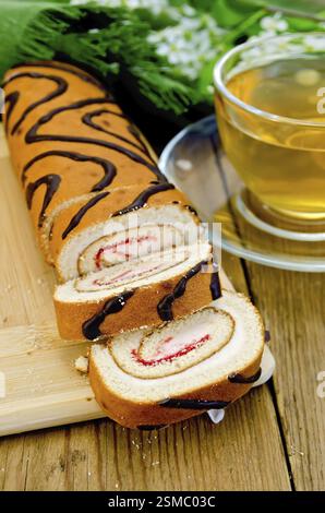 Biscuit roulade with cream and jam, a glass cup with tea, napkin, flowers and leaves of bird-cherry on a wooden board Stock Photo