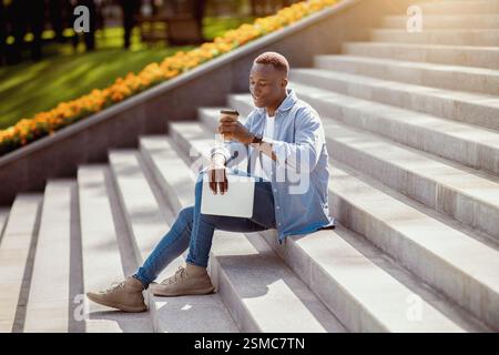 Positive black guy with tablet computer drinking takeaway coffee on stone stairs downtown Stock Photo