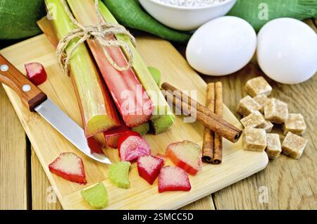 Bundle of stalks rhubarb, cut pieces of rhubarb, knife, sugar cubes, cinnamon, two eggs, flour, napkin on wooden board Stock Photo