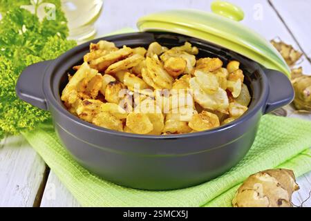 Jerusalem artichokes roasted in roasting pan with lid, fresh tubers on a napkin, parsley, vegetable oil on background of white wooden plank Stock Photo