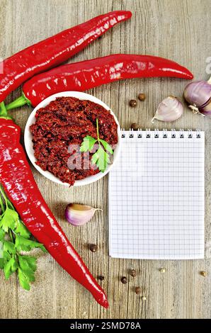 Tabasco in a white cup, fresh red peppers, garlic, peppercorns, parsley, notepad on an old wooden board Stock Photo