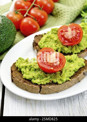 Two slices of rye bread with guakomole and tomato on a plate, napkin, avocado on a background of light wooden boards Stock Photo