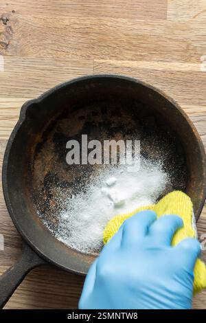 Person cleaning a rusty old cast iron frying pan using bicarbonate of soda and a scouring pad. On a wood board background. Stock Photo