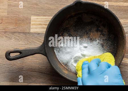 Person cleaning a rusty old cast iron frying pan using bicarbonate of soda and a scouring pad. On a wood board background. Stock Photo