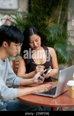 A beautiful, focused Asian woman attentively takes notes while collaborating with her colleague during a meeting at an outdoor table in a coffee shop, Stock Photo