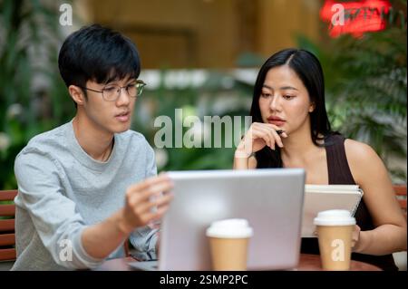 An attractive Asian woman attentively listens to her colleague's ideas, looking at a laptop screen while discussing work together at an outdoor table Stock Photo