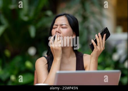 A tired Asian woman is feeling sleepy after a long day of working, yawning while sitting in front of her laptop and working in her apartment courtyard Stock Photo