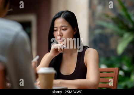 A beautiful, serious Asian woman focuses on reading something on her laptop while having an informal meeting with her colleague at a cafe. Stock Photo