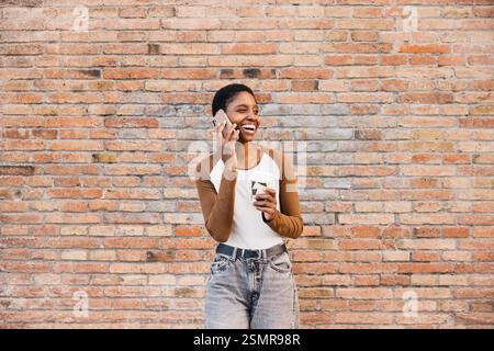Happy woman enjoying a coffee break, talking on smartphone against urban brick wall background Stock Photo