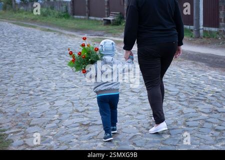 This image captures a heartwarming scene of a toddler carrying a bouquet of red flowers, holding hands with an adult. They are strolling down a quaint Stock Photo