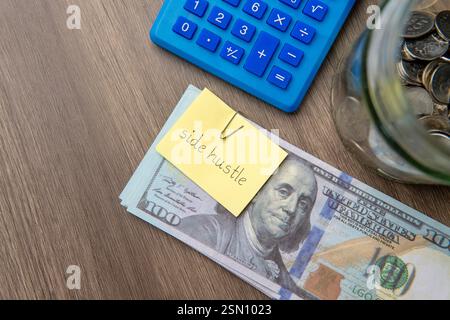 A sticky note with the words "side hustle" on top of a stack of US dollar bills, with a calculator and a jar of coins. Stock Photo