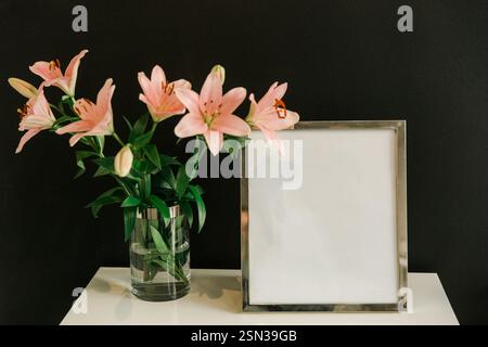 Bouquet of pink lilies in a glass vase on the table and empty frame Stock Photo