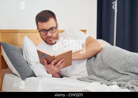 Man in white shirt and glasses laying down on the bed and reading book. Stock Photo