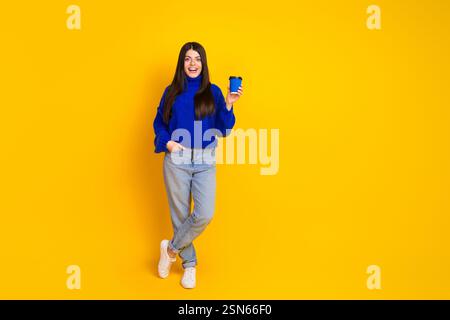 Smiling young woman in a blue knit sweater holding a coffee cup against a vibrant yellow background Stock Photo