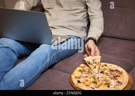 man sitting on sofa and eating pizza while working on laptop, close-up Stock Photo
