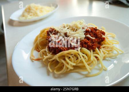 A dish of spaghetti pasta with minced meat in tomato sauce sprinkled with yellow cheese served on a white round big plate stands on the kitchen counte Stock Photo