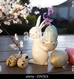 souvenir porcelain Easter bunny stands on the table, quail and chicken eggs nearby, spring white flowering tree branches. Home decor, festive Easter, Stock Photo
