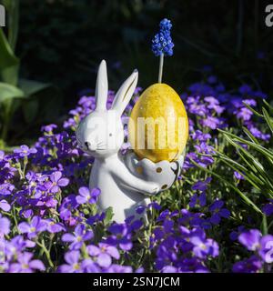 souvenir porcelain easter bunny with yellow painted egg among many blue - purple small flowers on flowerbed. beautiful festive easter atmosphere. Happ Stock Photo