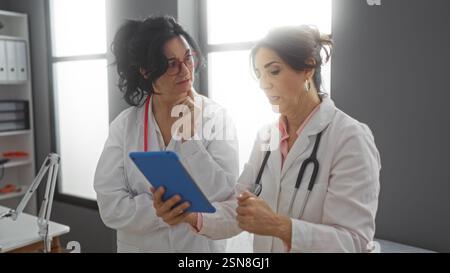 Two female doctors discussing patient data on a tablet in a modern medical office with natural lighting and professional decor, emphasizing collaborat Stock Photo