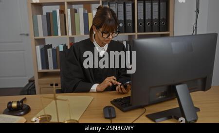 Female judge in black robe working at desk in office with books and files while looking at her phone Stock Photo