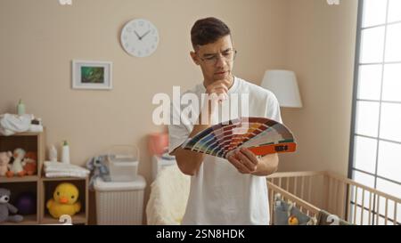 Young man in a clinic room examines color swatches, thoughtfully planning decor near a baby crib with nursery toys. Stock Photo