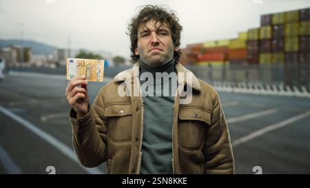 Man holding euro banknote stands confidently outdoors near the seaside port, with shipping containers and boats visible in the background, showcasing Stock Photo