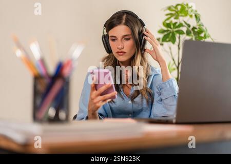 A focused young woman wearing headphones and using a smartphone while working on her laptop at a desk. The blurred foreground elements create depth, e Stock Photo
