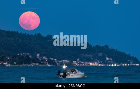 A boat is floating on the water with a full moon in the background. The moon is bright and pink, creating a serene and peaceful atmosphere. The boat i Stock Photo