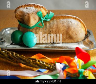 Easter lamb cake with a green ribbon around its neck and painted eggs on a silver platter, an Easter whip with colorful ribbons in the foreground. Stock Photo