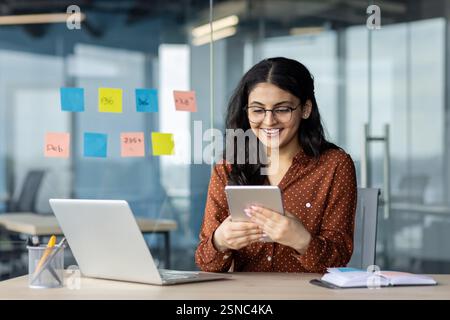 Successful businesswoman using tablet computer while sitting at desk with laptop in office. Female consultant employee of company happy and smiling. Stock Photo