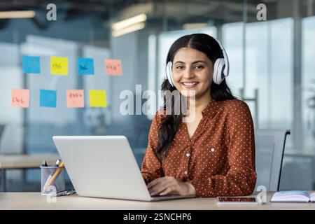 Young woman working inside office wearing headphones, programmer smiling and looking at camera, employee coding new software, developing applications. Stock Photo