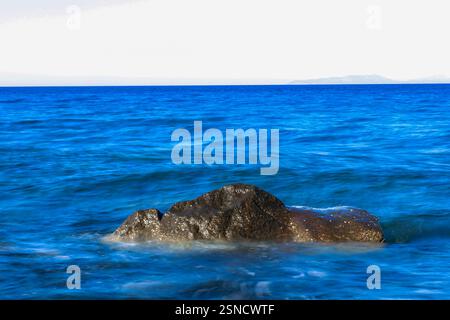dark, textured rock emerges from the smooth, flowing motion of the deep blue ocean Stock Photo