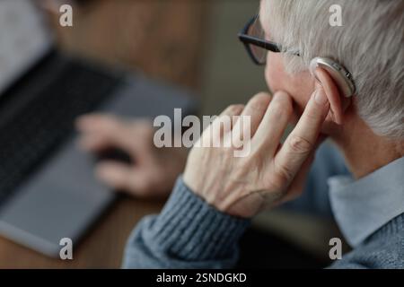 Close up on elderly mans hand tuning up hearing aid behind ear by pressing finger on device controlling volume of sound while working on laptop at home, assistive technology use, copy space Stock Photo