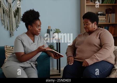 Smiling African American female social worker explaining elderly woman how to pay bills online on digital tablet pointing at computer screen, while sitting together in living room Stock Photo