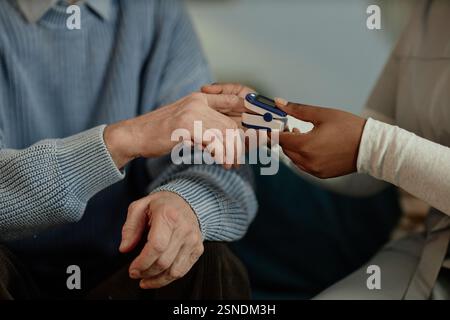 Cropped shot of African American female social worker checking up on elderly mans blood oxygen saturation using oximeter during home care visit, image in warm tones Stock Photo