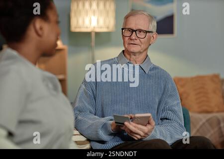 Medium portrait of white haired elderly man making electronic payment online on mobile phone while social worker assisting pensioner at home Stock Photo