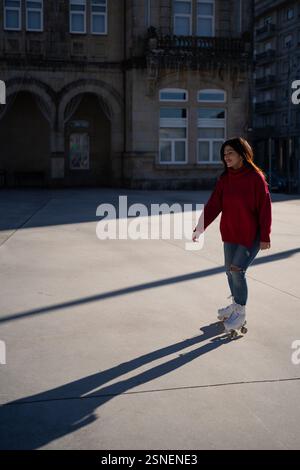 Young woman enjoys roller skating in a paved urban area, casting a long shadow on the ground while wearing a red turtleneck sweater and ripped jeans Stock Photo