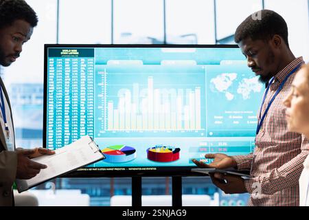 Team of workers presenting infographics and sales numbers on an interactive board, boardroom activity. Diverse group of professionals engages in a project management meeting for growth. Stock Photo