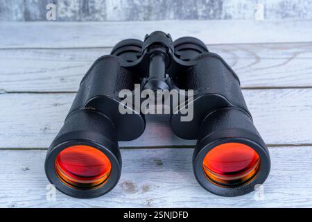Close-up of black binoculars with red-tinted lenses placed on a white wooden surface, symbolizing exploration, vision, and outdoor adventure. Stock Photo