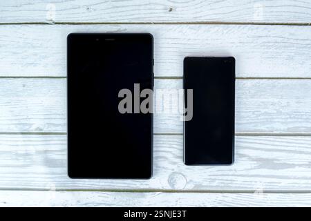Black tablet and smartphone with turned-off screens placed side by side on a white wooden surface. Top-down view. Stock Photo
