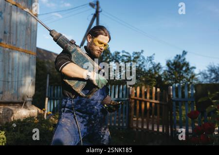 Man using a large drill outdoors in a garden area with clear blue sky and wooden fence Stock Photo