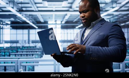 Computer scientist in server hub checking recovery plan on laptop, monitoring hardware energy consumption. IT specialist uses notebook in data center facility, ensuring rigs are functioning, camera B Stock Photo