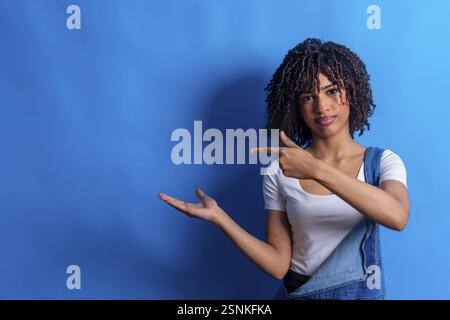 Young woman pointing her finger to the side and showing something with her open hand on a vibrant blue background Stock Photo